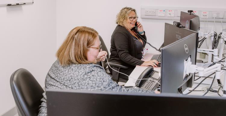Two women sitting in an office in front of computer monitors. One is on the phone.