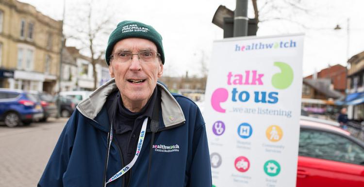 Older volunteer standing infront of a Healthwatch banner at an event