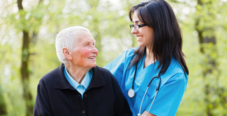 older patient with female nurse standing outside in front of trees