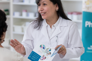 A pharmacist holding a leaflet, talking to a person in a pharmacy.