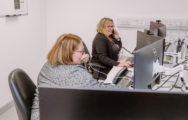 Two women sitting in an office in front of computer monitors. One is on the phone.